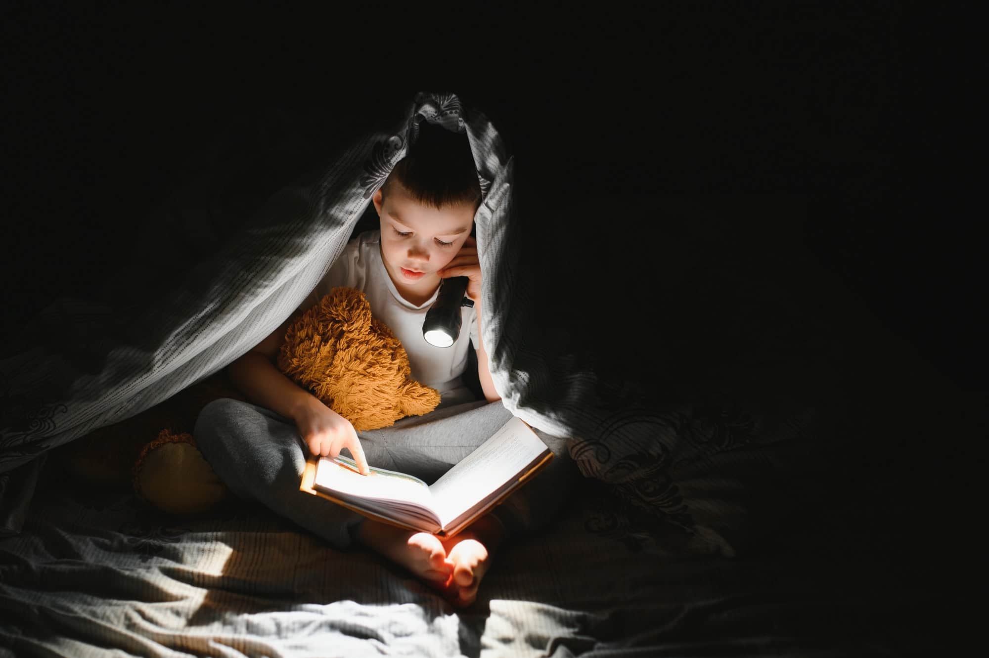 Boy with flashlight reading book under blanket at home.